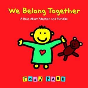 A colorful cover of the book 'We Belong Together: A Book About Adoption and Families' with a drawing of a child with yellow skin and a green shirt with a red heart in the center holding a brown teddy bear.