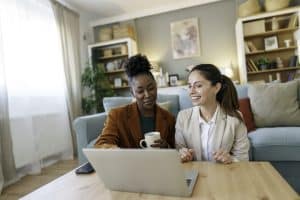 A black woman and her Caucasian colleague smile as they work on a laptop in a well-lit living room, exemplifying a home-based small business environment.